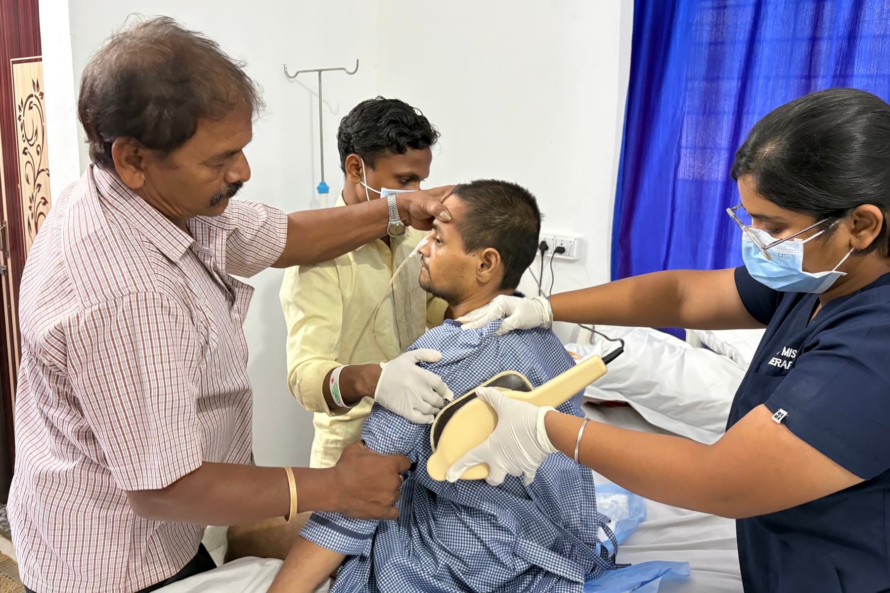 A DSW therapist gives a patient chest physiotherapy as he sits up in bed, with his family steadying him.